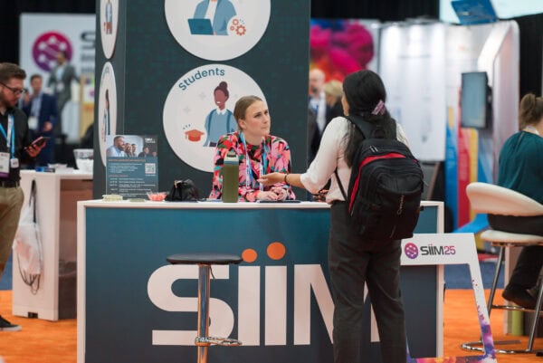 Professional woman at SIIM Annual Meeting booth engaging with attendee, showcasing event registration and educational programs for medical imaging and informatics professionals.