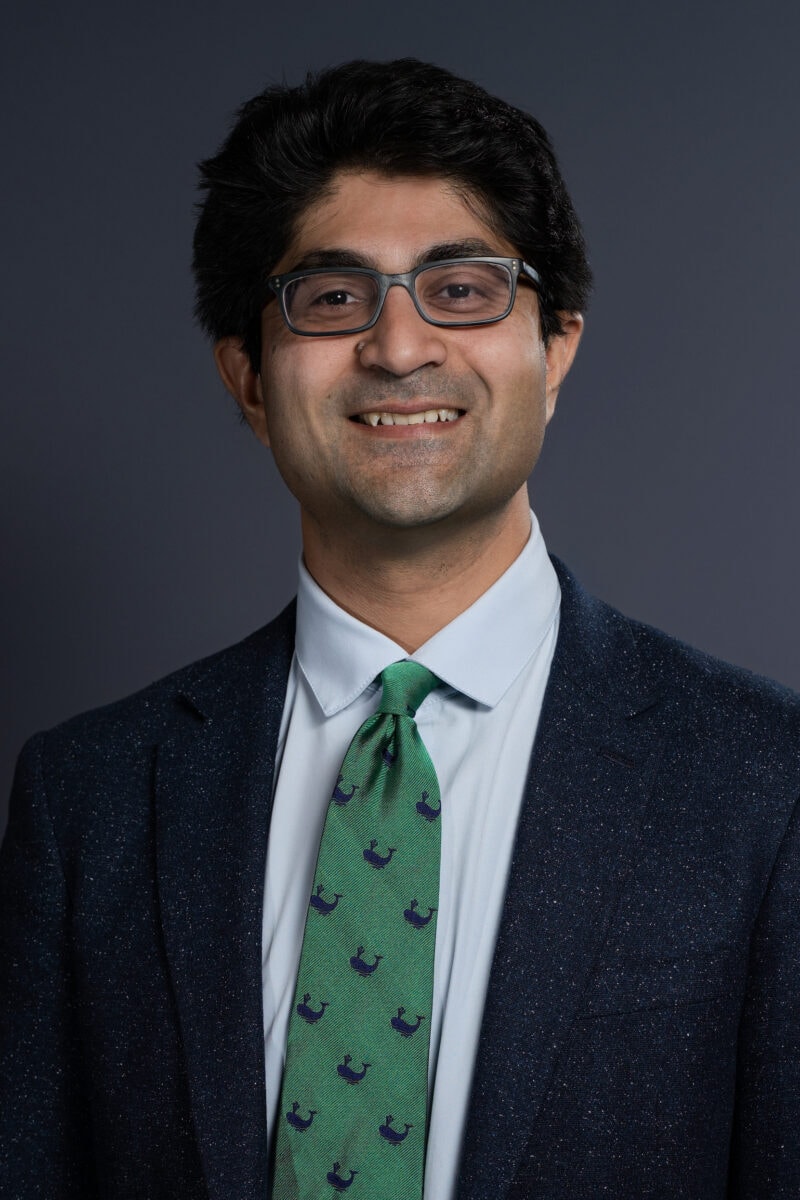 A professional portrait of a smiling man wearing glasses, a dark blazer, white shirt, and a green tie with navy blue patterns, set against a plain dark background.