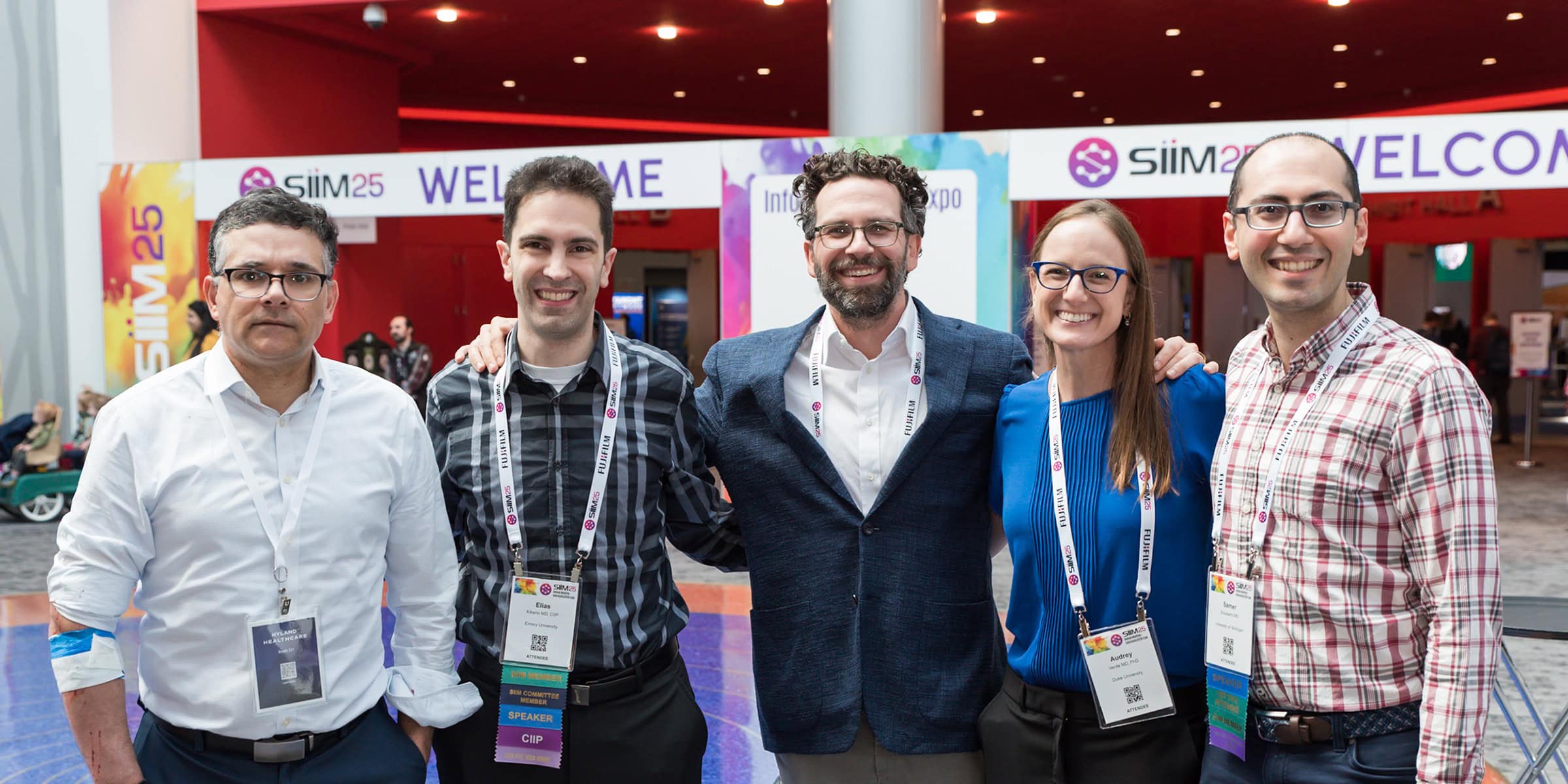 1. Group of diverse medical professionals and conference attendees smiling at the SIIM Annual Meeting in front of exhibitor banners.