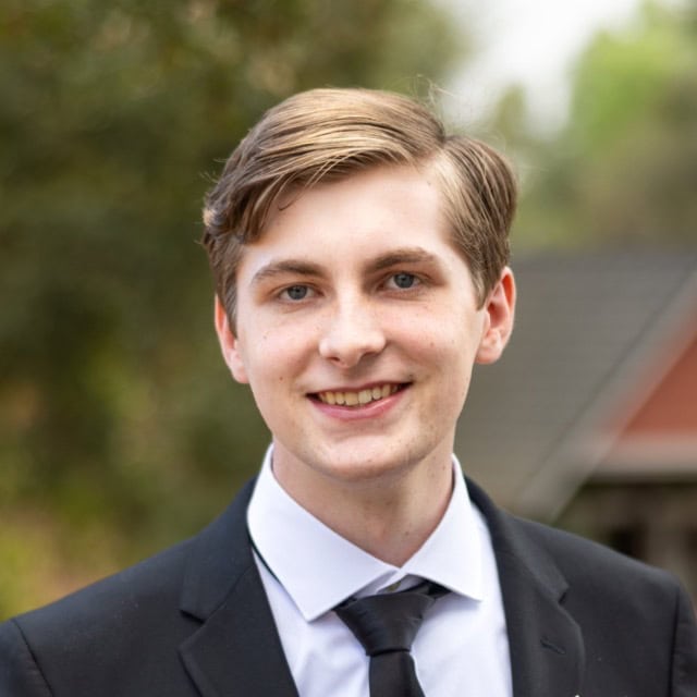 Young male medical professional smiling at SIIM Annual Meeting, wearing formal suit, with outdoor background.