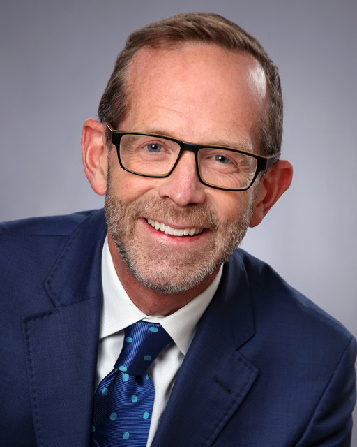 An image of a smiling professional man wearing glasses, a navy suit, and a blue tie, attending the SIIM Annual Meeting, a leading event in medical imaging and informatics.