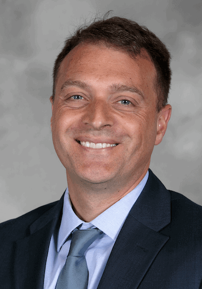 1. Professional male at SIIM Annual Meeting wearing a suit, smiling, with a gray background; radiology imaging, medical innovation, healthcare technology.