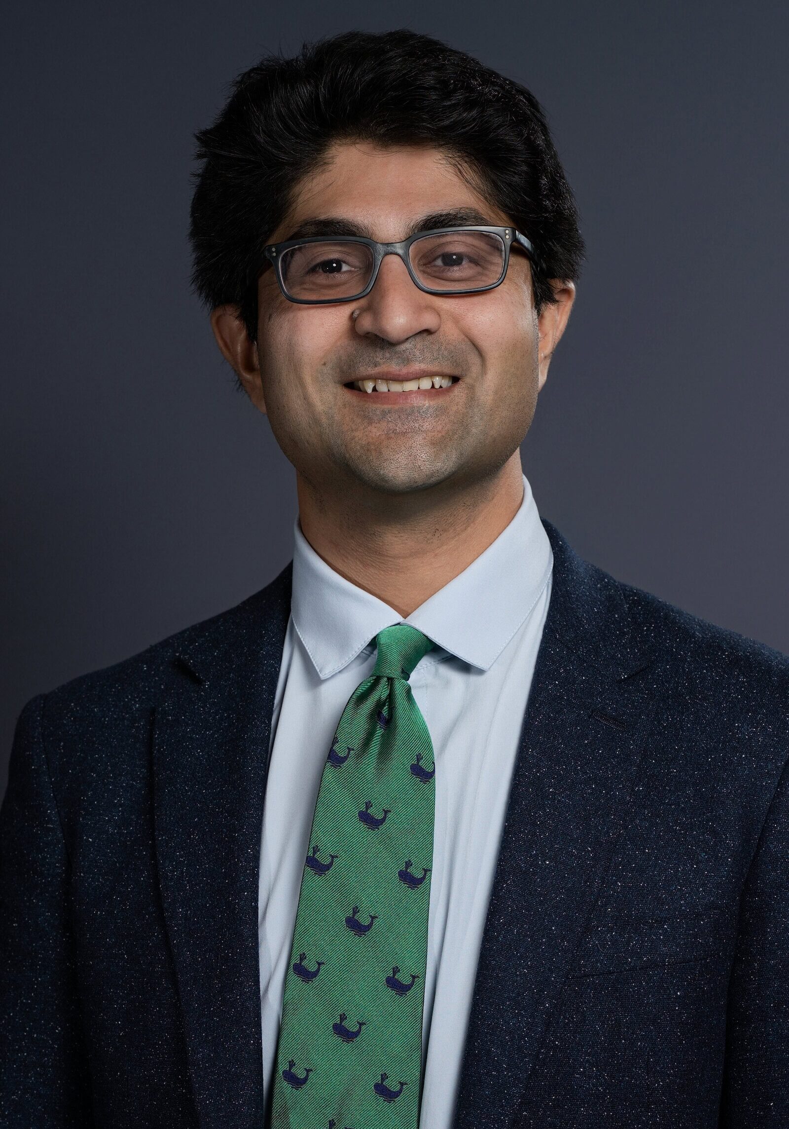 A professional portrait of a smiling man wearing glasses, a dark blazer, white shirt, and a green tie with navy blue patterns, set against a plain dark background.